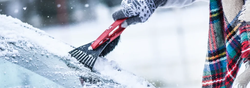 Eine Person reinigt Schnee von einem Autofenster mit einem roten Schneeschieber, umgeben von winterlicher Landschaft.