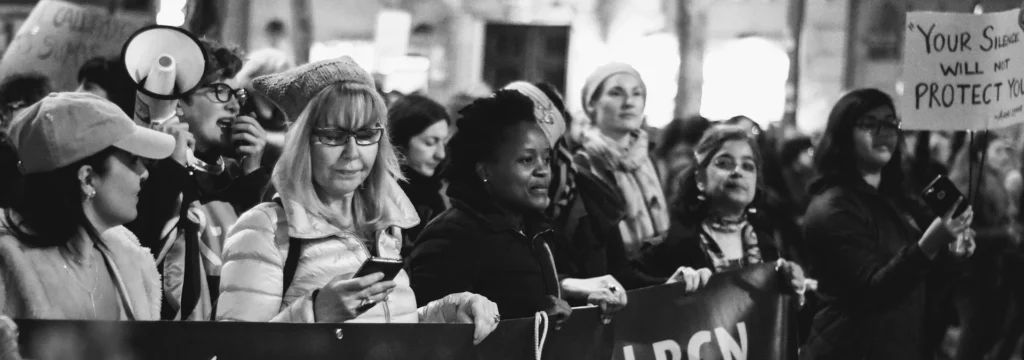 Gruppe von Frauen bei einer Demonstration, einige halten Schilder und andere nutzen Smartphones.