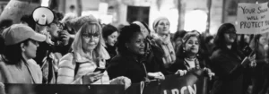 Gruppe von Frauen bei einer Demonstration, einige halten Schilder und andere nutzen Smartphones.