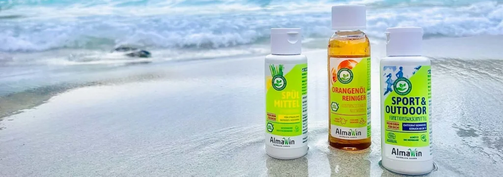 Drei Flaschen Almawin Reinigungsprodukte auf sandigem Strand mit Wasser im Hintergrund.