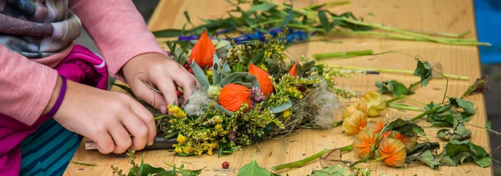 Hände eines Kindes, die einen Frühlingskranz aus verschiedenen Blumen und Blättern auf einem Tisch gestalten.