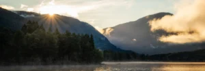 Sonnenaufgang hinter Bergen mit Lichtstrahlen und bewaldeter Landschaft am Wasser.